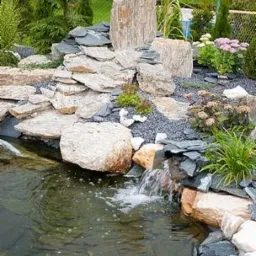 Oczko wodne z kaskadą i kamienną dekoracją w Brzeszczach. Ujęcie z bliska na wodę spływającą po kamieniach. Otoczenie obsadzone roślinami.