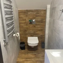 Wnętrze łazienki z drewnopodobną podłogą i ścianą za toaletą, szara ściana z grzejnikiem drabinkowym, wisząca toaleta z chromowanym przyciskiem spłukującym, czarny kosz na śmieci i szara szczotka...