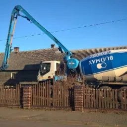 Betonomieszarka Budmax z wysięgnikiem podającym beton na budowę domu z niebieskim dachem w tle.