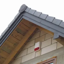 Zewnętrzna czujka alarmowa z czerwoną diodą zamontowana na ścianie budynku z bloczków, widoczna spod okapu dachu pokrytego szarą dachówką.