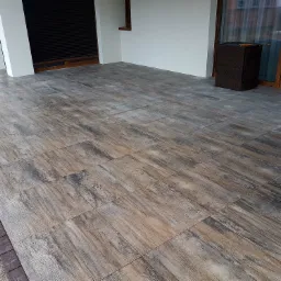 Wyłożony płytkami taras z efektem drewna, obrzeże z ciemnej kostki, widoczne białe ściany budynku i brązowe drzwi tarasowe.