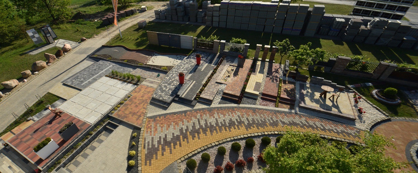 Ekspozycja różnorodnych wzorów kostki brukowej i granitowej na zewnątrz, z paletami kostki w tle. Widok z góry prezentuje bogactwo kolorów i faktur.