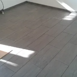 Ukończona podłoga z szarych, prostokątnych płytek, z widocznym fragmentem płyty z boku. Świeże, jasne wnętrze z listwami przypodłogowymi.