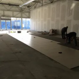 Układanie jasnych, kwadratowych płytek na podłodze w dużym pomieszczeniu. Dwóch pracowników w trakcie pracy. Ściany przygotowane pod malowanie. Widoczne narzędzia i materiały.