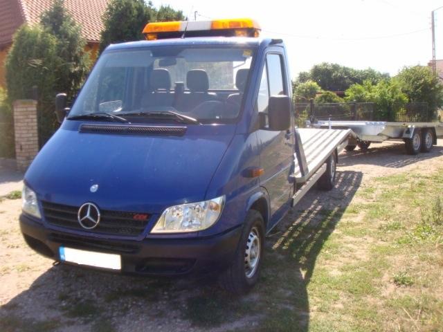 Niebieska laweta Mercedes z aluminiową platformą i przyczepą na trawiastym terenie. Pomarańczowe światła ostrzegawcze na dachu.