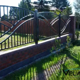 Eleganckie ogrodzenie kute z ceglaną podmurówką i falistym wzorem na tle zielonego trawnika i drewnianego płotu. Widoczne słupki z cegły.