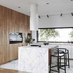 INTUS GROUP - Nowoczesna kuchnia z marmurową wyspą, drewnianymi szafkami i minimalistycznym oświetleniem punktowym, widok na ogród przez duże okno z żaluzjami.