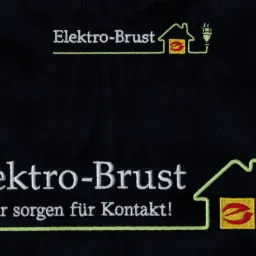 Ciemna tkanina z wyhaftowanym logo firmy Elektro-Brust, przedstawiającym dom z wtyczką i hasłem '...wir sorgen für Kontakt!'
