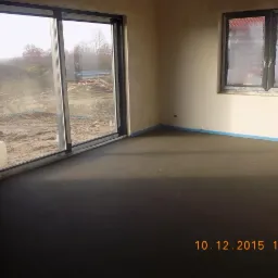 Świeżo wylana, ciemna posadzka w jasnym pomieszczeniu z dużym oknem i widokiem na plac budowy. Niebieska taśma zabezpieczająca przy ścianach.