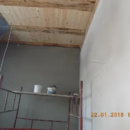 Ściana po tynkowaniu z widocznym rusztowaniem, wiaderkami z zaprawą i drewnianym sufitem. Świeży tynk na ścianie, data wykonania widoczna w prawym dolnym rogu.