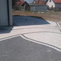 Ułożona nawierzchnia z kostki brukowej w odcieniach szarości i bieli, z dekoracyjnymi liniami oddzielającymi poszczególne sekcje, widok z perspektywy ogrodzenia na posesję z garażem i domami w tle.