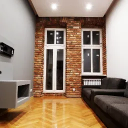 Nowoczesny salon z ceglaną ścianą, białymi oknami i szarą zabudową TV z uchwytem. Parkiet w jodełkę i szara sofa dopełniają wnętrze.