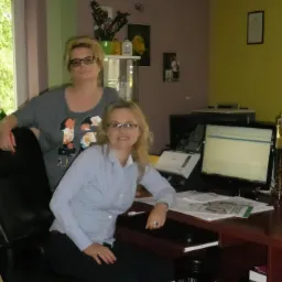 Dwie kobiety w biurze. Jedna siedzi przy biurku z komputerem, druga stoi za nią. Na biurku dokumenty i lampka. W tle okno i kwiaty.