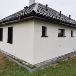 Nowo otynkowany, biały dom jednorodzinny z czarną dachówką i ramami okiennymi, widok z zewnątrz, świeżo położona kostka obok budynku.