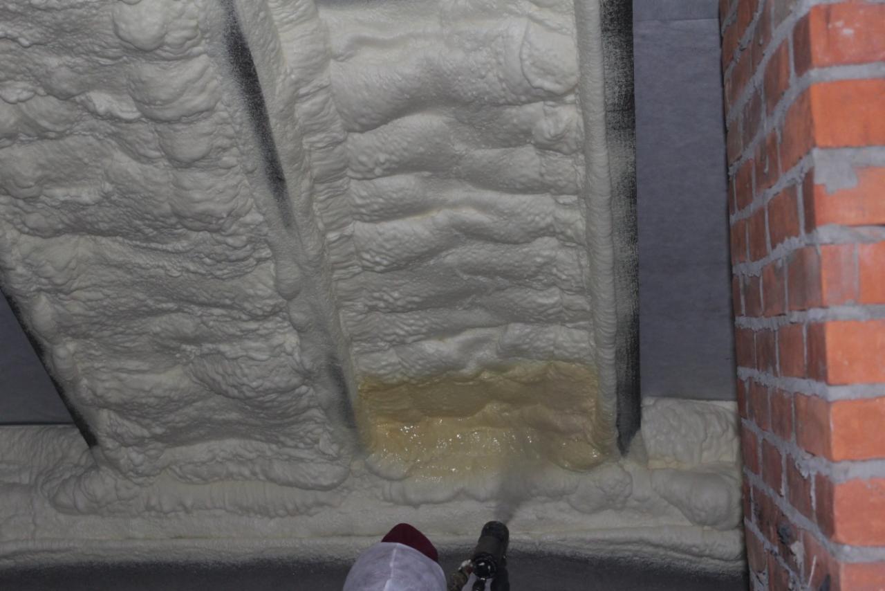 Aplikacja pianki izolacyjnej na stropie budynku, widoczna warstwa mokrej piany i fragment muru z cegły.