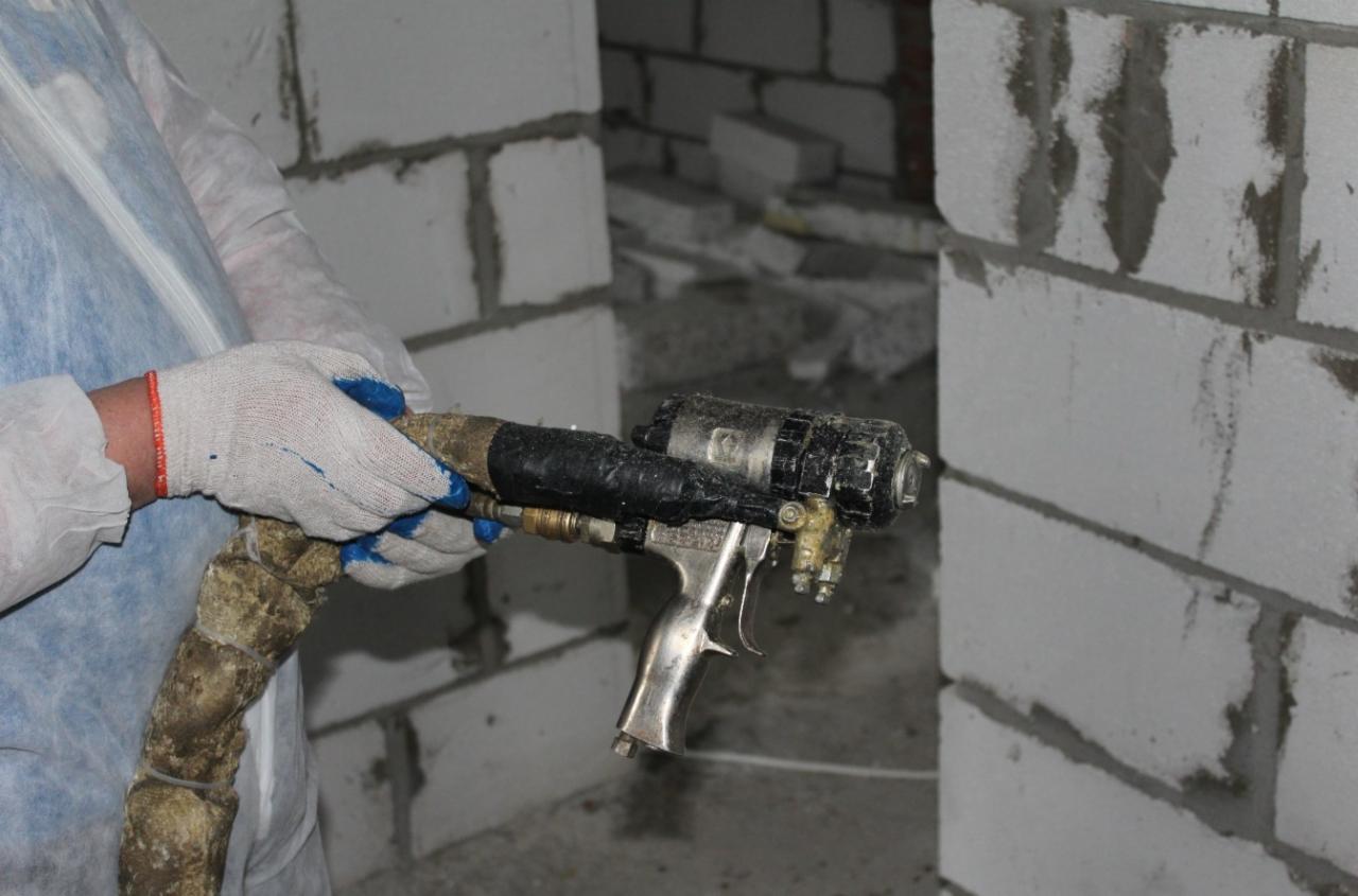 Pracownik w kombinezonie ochronnym i rękawicach trzyma pistolet natryskowy do izolacji piankowej na tle surowych ścian z białego betonu komórkowego.