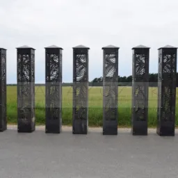 Siedem metalowych słupków ogrodzeniowych z wycinanymi wzorami liści palmowych, ustawionych w rzędzie na asfalcie, z polem i lasem w tle.