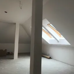 Poddasze w trakcie wykańczania, widoczne okna dachowe, słup konstrukcyjny, ściany przygotowane do malowania, widoczne puszki elektryczne, na podłodze zwinięta rolka materiału zabezpieczającego.