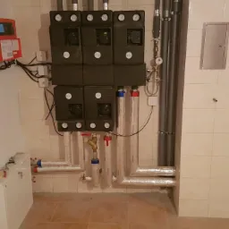 Złożony system instalacji grzewczej z kotłem, pompami obiegowymi i sterownikiem w pomieszczeniu wyłożonym płytkami ceramicznymi; widoczne izolowane rury, manometry i przewody elektryczne.