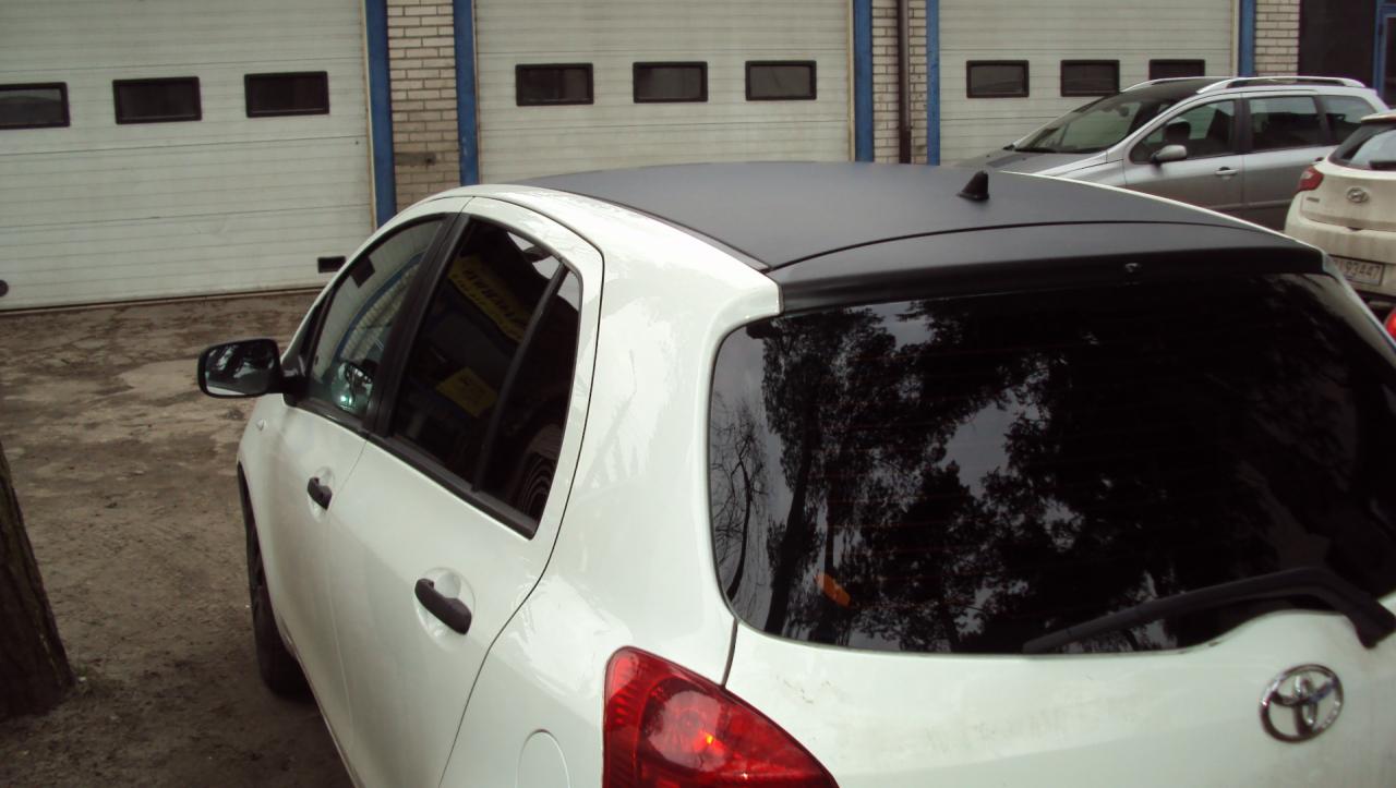 Biały samochód z czarnym dachem po tuningu. Widoczne tylne światła i czarna szyba. W tle brama garażowa i inne auta.