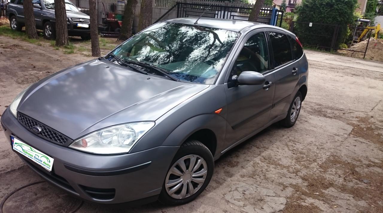 Szary Ford Focus hatchback na zewnątrz z widocznym odbiciem drzew na karoserii. W tle inne samochody i ogrodzenie posesji.