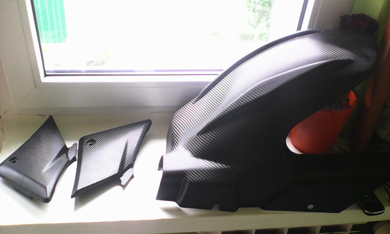 Elementy karoserii motocykla po tuningu, pokryte folią imitującą karbon, leżące na parapecie przy oknie. Widoczne detale faktury materiału.