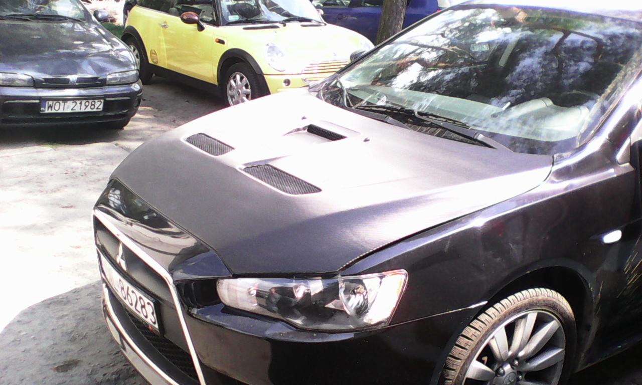 Czarny samochód z włókna węglowego na masce i wlotami powietrza, tuning wizualny, inne auta w tle.