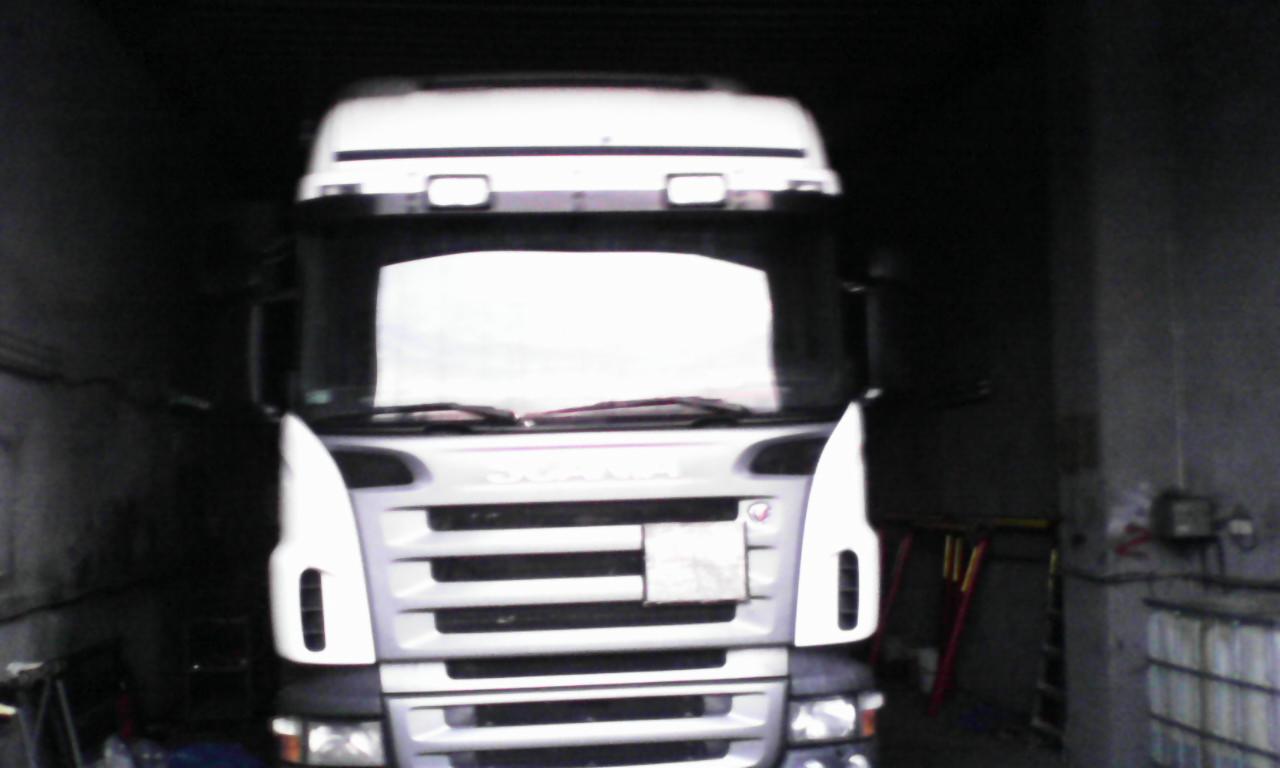 Biała ciężarówka SCANIA w ciemnym pomieszczeniu, widok z przodu. Na grillu pojazdu widoczna prostokątna plama. Słabe oświetlenie, garażowe tło.