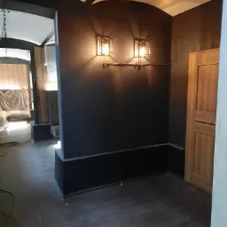 Wnętrze z ciemnymi ścianami, dwoma industrialnymi lampami ściennymi i drewnianymi drzwiami, widok na sąsiednie pomieszczenie z zasłoniętymi meblami.