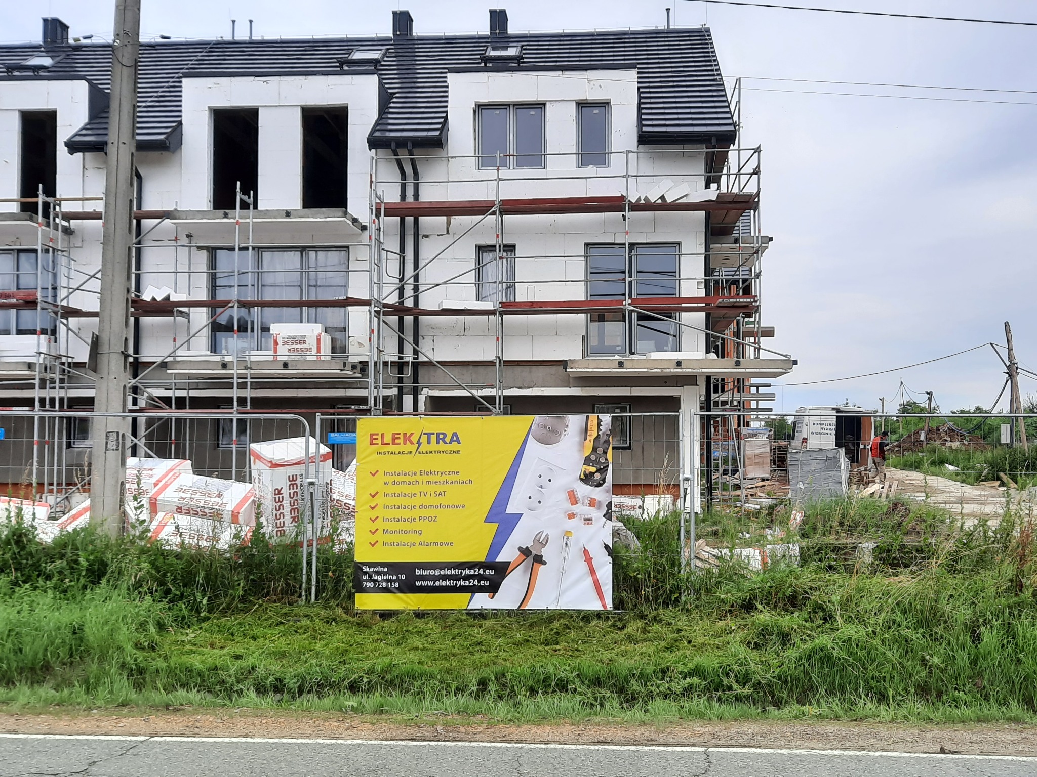 Nowo budowane domy w zabudowie szeregowej, w trakcie instalacji, widoczne rusztowania i materiały budowlane, a na pierwszym planie baner reklamowy firmy oferującej usługi elektryczne i instalacyjne.