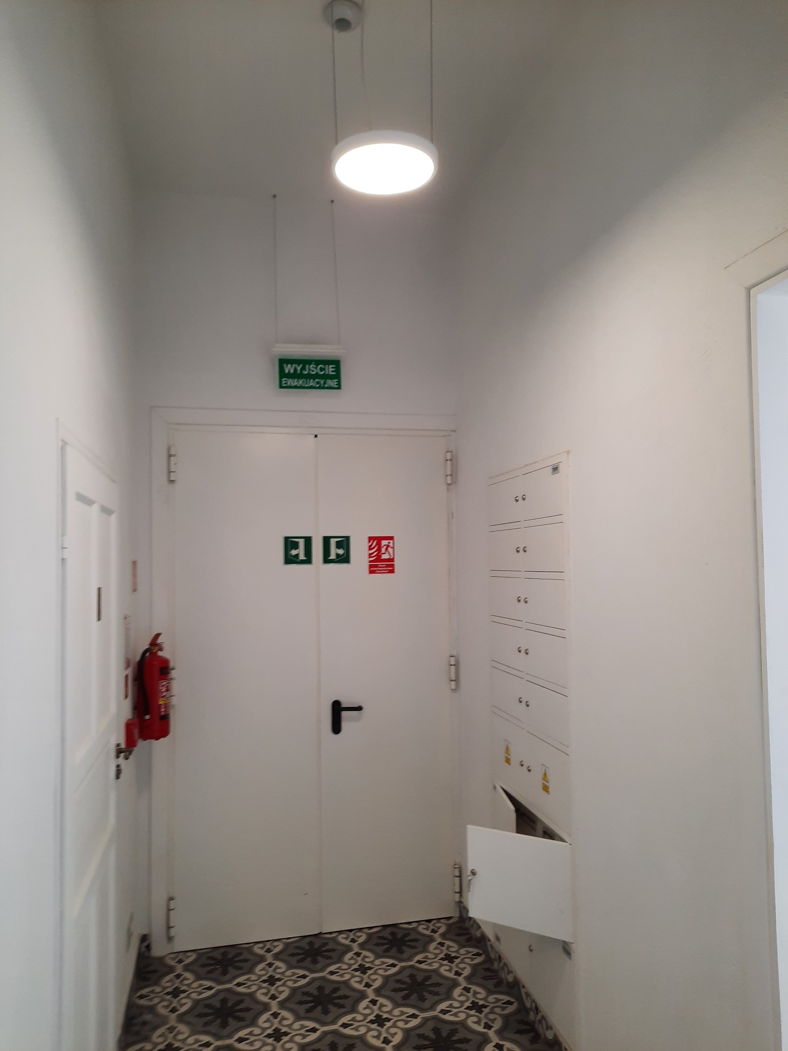 Korytarz z białymi ścianami, wyjściem ewakuacyjnym z gaśnicą, szafką elektryczną z otwartymi drzwiczkami i wzorzystą podłogą.