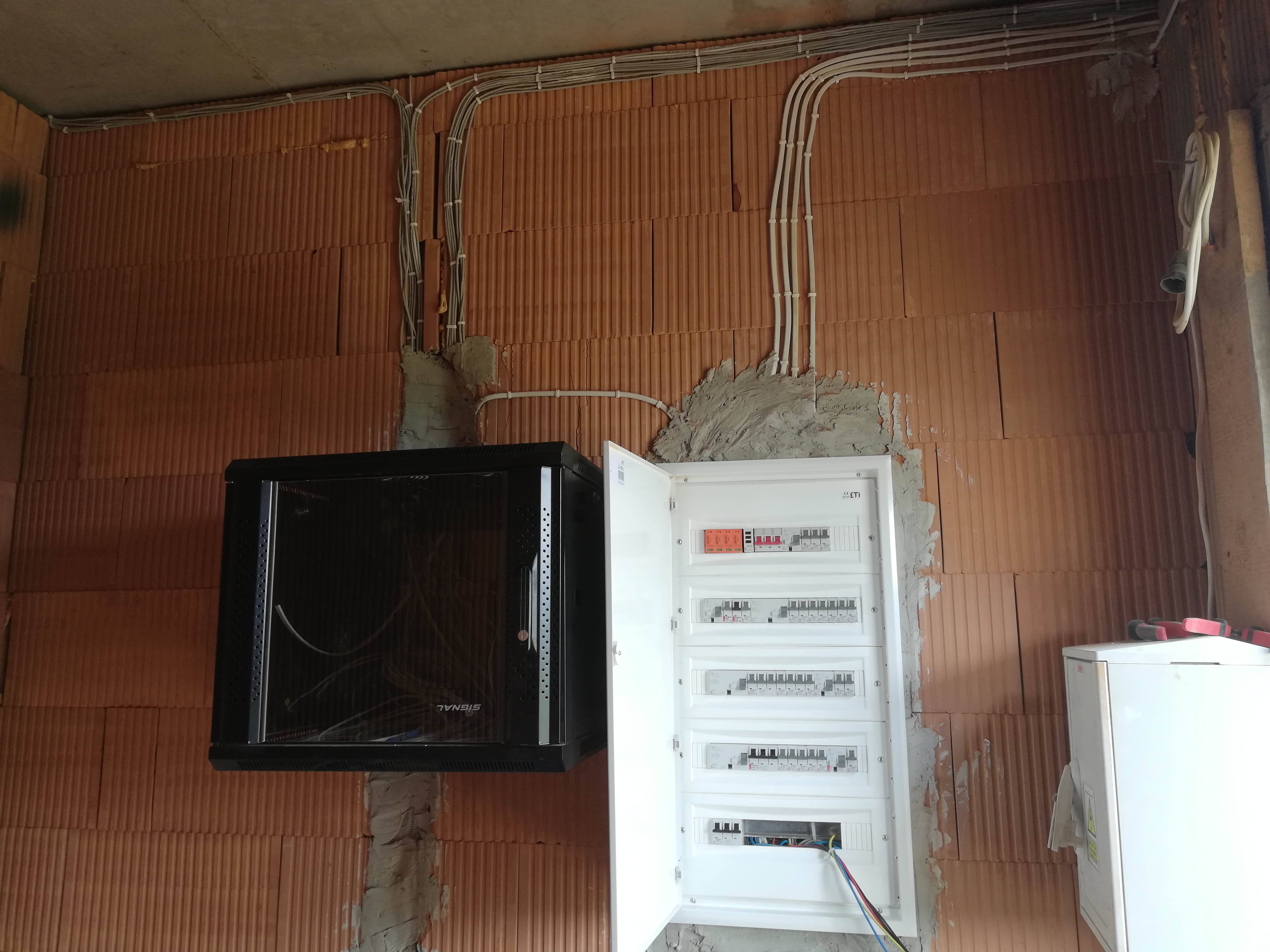 Rozdzielnica elektryczna z otwartymi drzwiczkami, zamontowana na ceglanej ścianie, z widocznymi kablami w peszlach biegnącymi po ścianie i szafa rack obok, w surowym stanie wykończenia.
