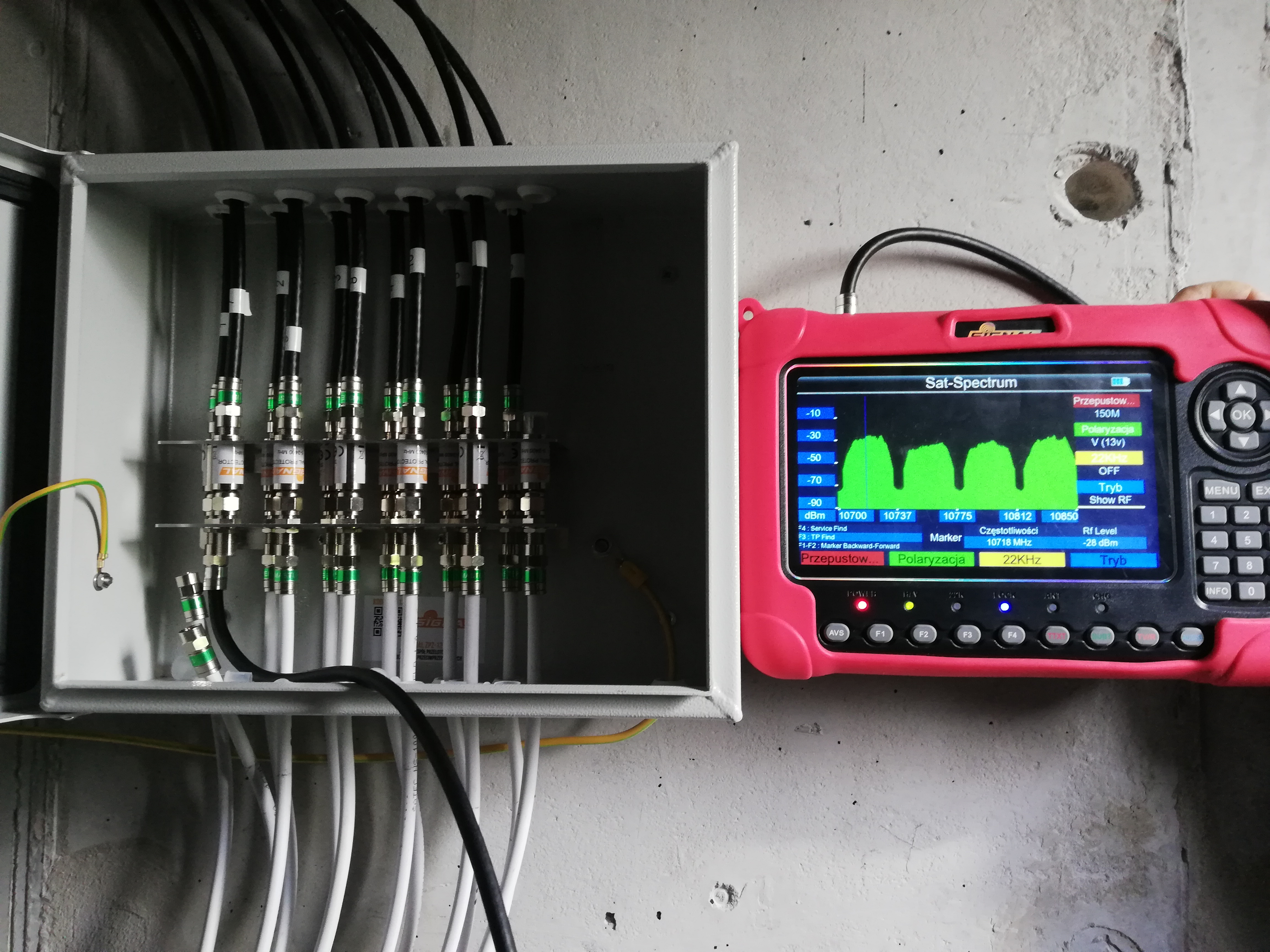 Rozdzielnica z kablami koncentrycznymi i miernik sygnału satelitarnego w obudowie, wyświetlający spektrum częstotliwości. Widoczne złącza F, przewody zasilające, uziemienie i elementy instalacji...
