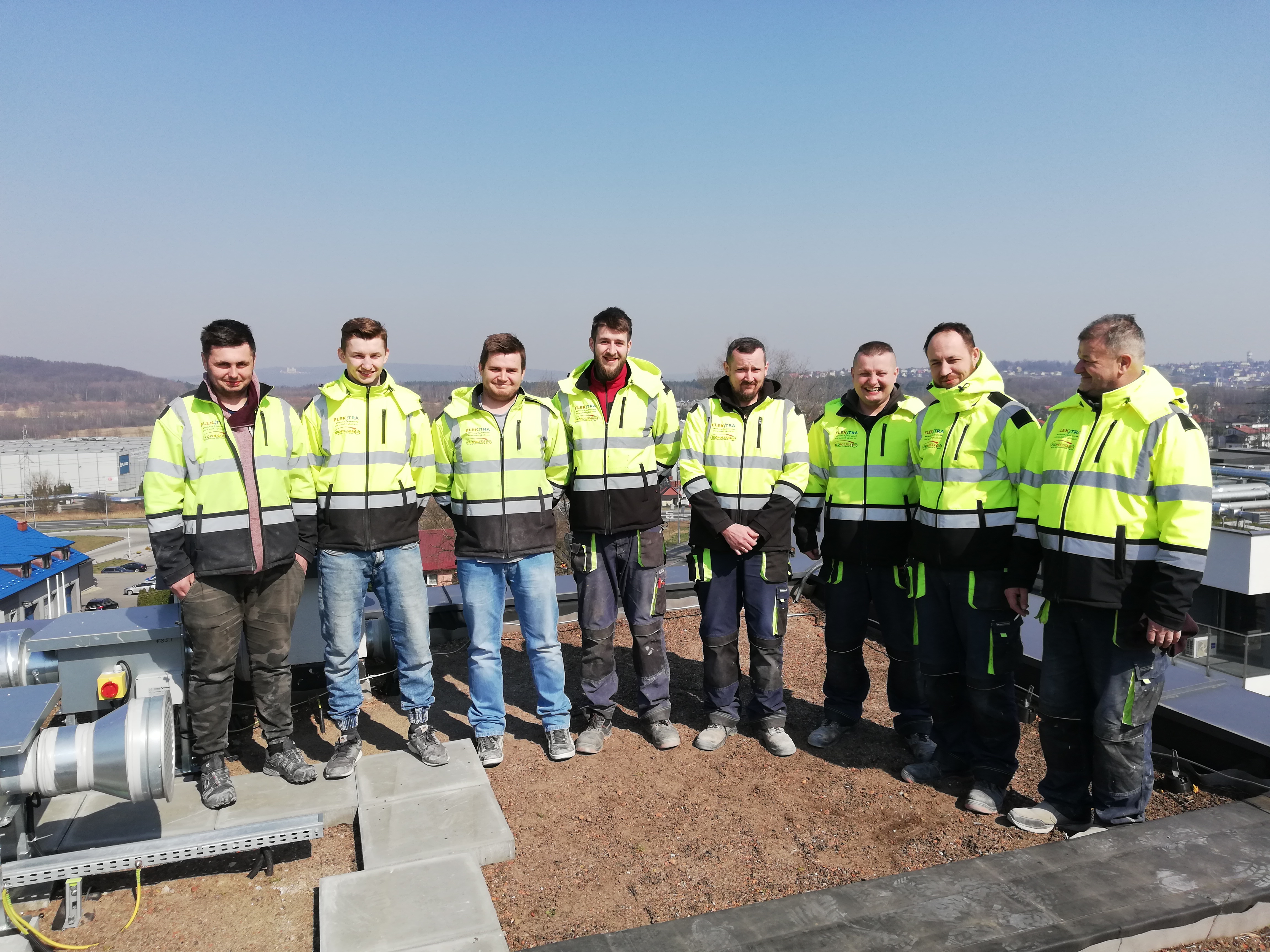 Grupa ośmiu elektryków w odblaskowych kurtkach firmy Elektra Troposka pozuje na dachu budynku z instalacją wentylacyjną, z widokiem na zabudowania miejskie w tle.