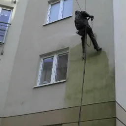 Pracownik na linach myje elewację budynku, usuwając zabrudzenia. Widoczny fragment umytej i brudnej ściany. Kąt ujęcia z dołu, na tle okien budynku.