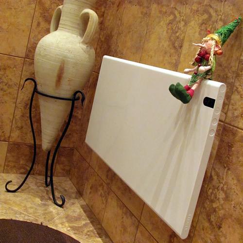 Biały, płaski grzejnik elektryczny na ścianie z brązowych płytek, obok stoi ozdobna amfora na metalowym stojaku, na grzejniku siedzi figurka elfa.