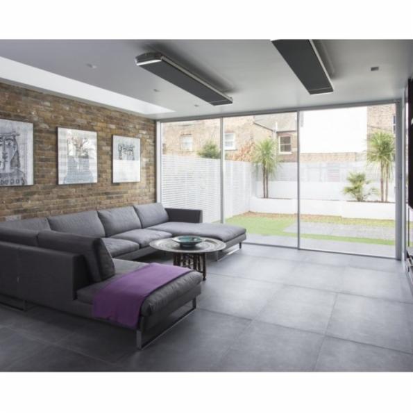 Nowoczesny salon z szarą sofą narożną, ceglaną ścianą z grafikami i dwoma podłużnymi panelami grzewczymi zamontowanymi na suficie, widok na ogród przez duże okno.