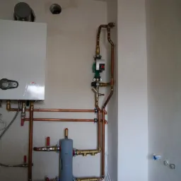 Biały, wiszący piec gazowy z podłączonymi miedzianymi rurami, pompą obiegową i szarym zbiornikiem wyrównawczym na tle surowej ściany w trakcie wykańczania.