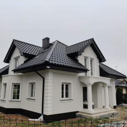 BUDMAX - Nowoczesny dom jednorodzinny z białą elewacją, czarnym dachem i kolumnami przy wejściu, widoczne detale sztukaterii i świeżo pomalowany cokół.