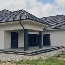 BUDMAX - Nowoczesny dom jednorodzinny z białą elewacją i ciemną dachówką, zadaszony taras wyłożony szarą kostką brukową, w tle niezagospodarowany teren z widoczną dziecięcą zabawką.