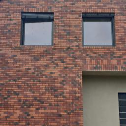 BUDMAX - Nowoczesny dom murowany z elewacją z czerwonej cegły klinkierowej z ciemnymi akcentami, czarnymi ramami okiennymi i fragmentem tynkowanej ściany w kolorze beżowym.