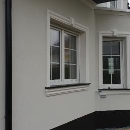 BUDMAX - Fragment elewacji budynku z dwoma oknami z białymi ramami i ozdobnymi gzymsami, wykończony jasnym tynkiem strukturalnym i czarnym pasem przy gruncie, widoczne elementy dekoracyjne w postaci pilastrów.