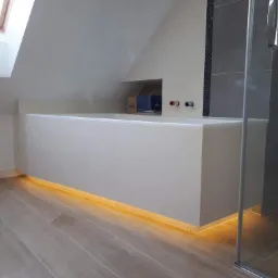 Nowoczesna łazienka na poddaszu z podświetlaną wanną obudowaną białymi płytkami, szara ściana z mozaiką i przeszklona kabina prysznicowa.