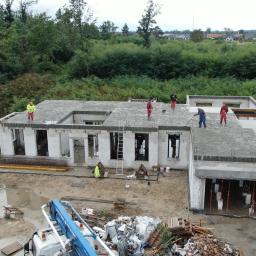 BUDMAX - Budowa domu jednorodzinnego w stanie surowym otwartym, widok z góry na ekipę pracującą na dachu, z użyciem długich łat, w tle zalesiony teren i zabudowania.