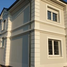 BUDMAX - Dwukondygnacyjny dom z jasną elewacją, dekoracyjnymi elementami architektonicznymi, balkonem z czarną balustradą i ciemnym dachem z dachówki.