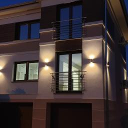 BUDMAX - Dwukondygnacyjny dom z jasną elewacją, oświetlony zewnętrznymi lampami ściennymi i liniowymi wbudowanymi w elewację, sfotografowany o zmierzchu.