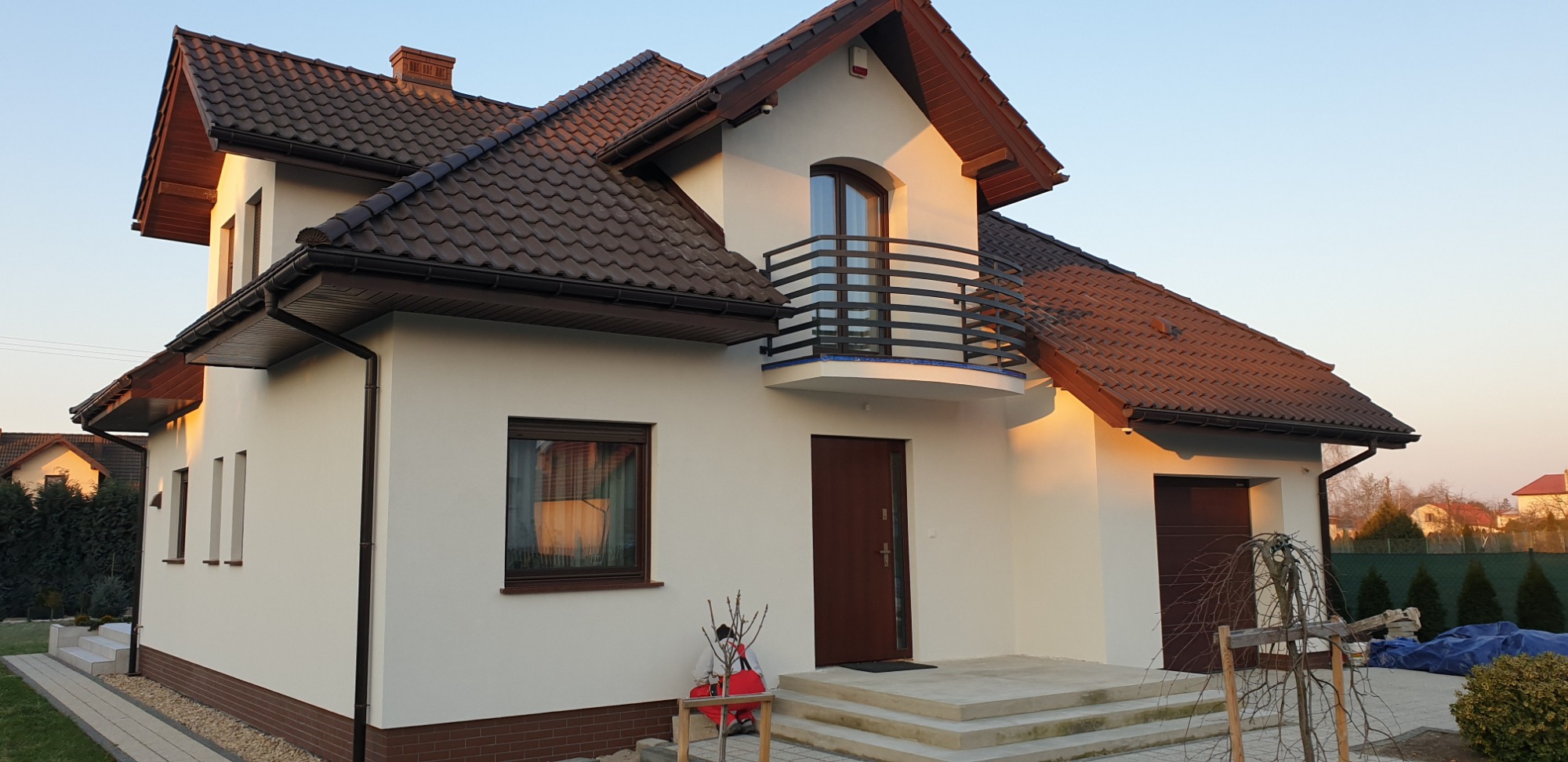 Nowo wybudowany, dwukondygnacyjny dom murowany z balkonem i brązową dachówką, oświetlony popołudniowym słońcem, z widocznymi elementami wykończenia elewacji.