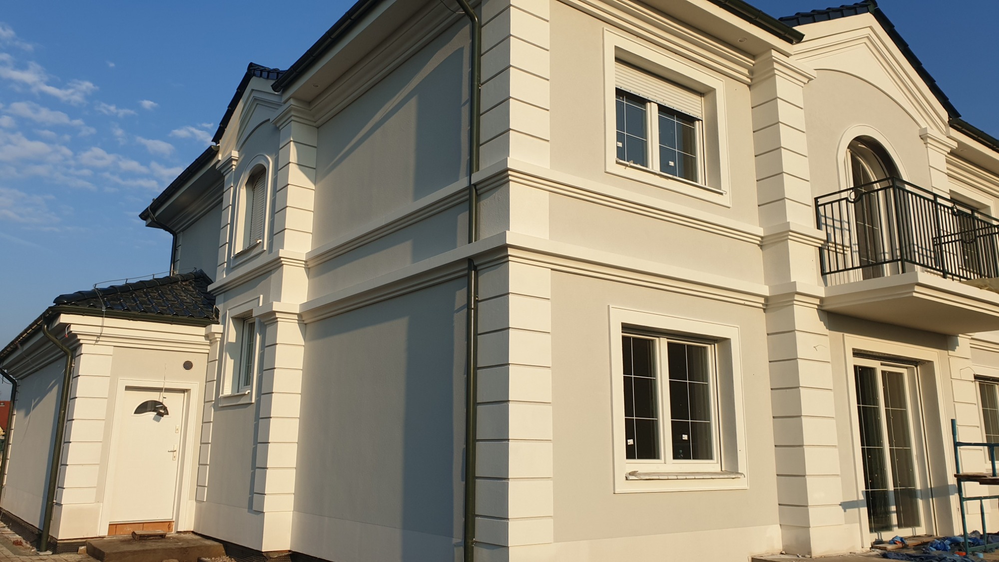 Dwukondygnacyjny dom z jasną elewacją, dekoracyjnymi elementami architektonicznymi, balkonem z czarną balustradą i ciemnym dachem z dachówki.
