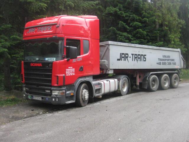 Czerwona ciężarówka Scania z szarą naczepą typu wywrotka firmy JAR-TRANS na asfalcie, z zielonym lasem w tle. Widoczny numer telefonu.