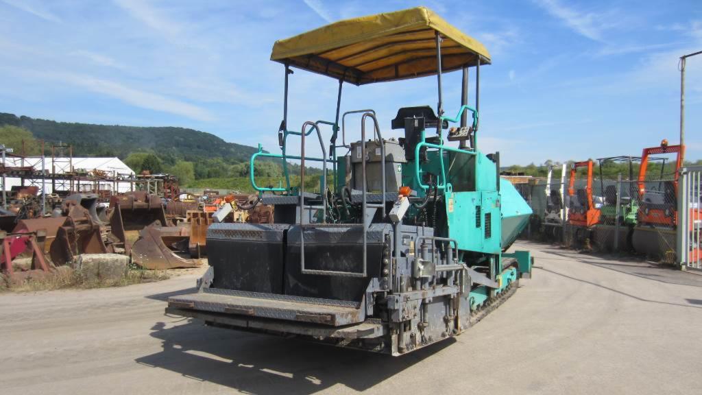 Rozściełacz VOGELE SUPER 1400 sfotografowany z tyłu, z widocznymi elementami konstrukcyjnymi i żółtym zadaszeniem, na placu z innymi maszynami budowlanymi i zielonymi wzgórzami w tle.